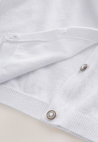 Close-up van een witte gebreide cardigan met twee zilverkleurige, met randjes afgewerkte knopen, dichtgemaakt nabij de geribbelde zoom.