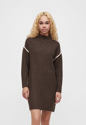 Femme aux longs cheveux blonds bouclés portant une robe en maille marron à manches longues avec des détails de passepoil blanc sur les manches.