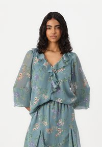 Blomstermønstret bluse i lys teal chiffon, med rynket halsringning og brede ærmer, med et integrert mønster av flerfargede blomster.