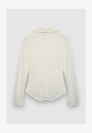 Chemise blanche texturée à manches longues avec col, poignets boutonnés et ourlet légèrement arrondi. Le motif subtil ajoute un intérêt visuel.