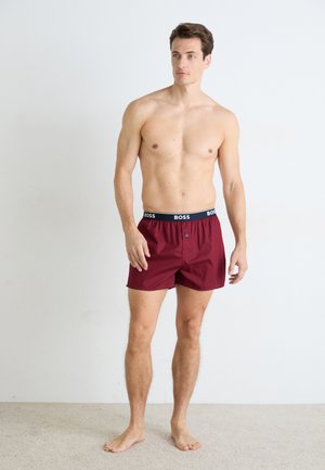 Homme torse nu, debout pieds nus sur un tapis clair, portant un short en maroon avec une ceinture marine portant l'inscription "BOSS", devant un mur blanc uni.