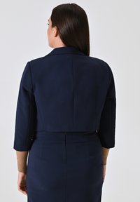 Korte marineblauwe jas met driekwartmouwen, getailleerde pasvorm en gestructureerde schouders. De achterkant heeft een nette naad en subtiele textuurelementen.