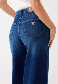 Jeans de mezclilla en azul oscuro con una cintura alta. Presentan una etiqueta de cuero en la parte trasera y una etiqueta con un logo en triángulo en el bolsillo. Costuras distintivas.