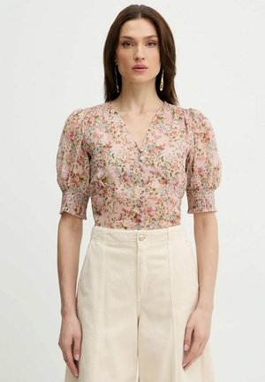 Donna che indossa una camicetta rosa a fiori con bottoni, maniche a sbuffo e pantaloni color crema a vita alta, su sfondo bianco uniforme.