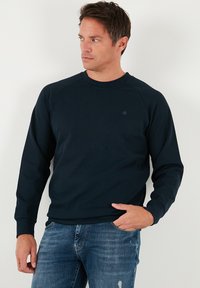 Tengerészkék pulóver kerek nyakkivágással és raglán ujjakkal. Kis logó látható a mellkasnál, valamint bordázott mandzsetták és szegélyek. Pamut keverék anyagból készült.