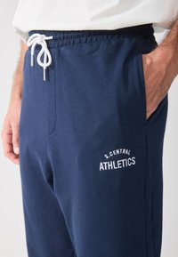 Marineblå bomullsbukser med strikk i livet, sidelommer og hvit brodert tekst som sier "S.CENTRAL ATHLETICS." Glatt tekstur.