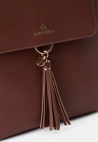 Bolsa de mão em pele castanha com um anel em tom dourado, decoração em forma de borlas e logótipo "Anna Field" em relevo na aba.