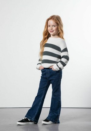 Młoda dziewczyna o długich rudych włosach, ubrana w biało-szary sweter w paski, niebieskie dżinsy i czarne tenisówki, stoi z rękami w kieszeniach.