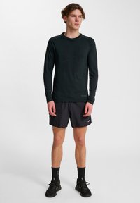 Zwart sportshirt met lange mouwen en een subtiel logo, gecombineerd met donkere shorts, gedragen met zwarte schoenen en sokken. Eenvoudig modern ontwerp.