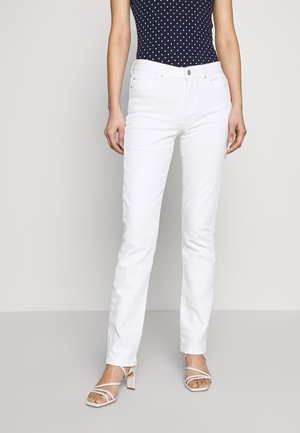 Nainen, jolla on päällään istuvat valkoiset farkut, laivastonsininen hihaton pilkullinen yläosa ja valkoiset remmikkäät blokki-korkokengät, seisoo yksiväristä taustaa vasten.