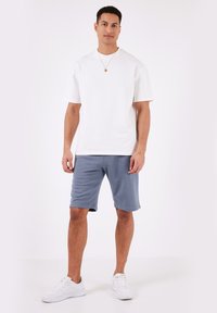 Biely bavlnený tričko s krátkymi rukávmi v kombinácii s modrými bavlnenými šortkami. Model má na sebe biele tenisky. Jednoduchý, ležérny outfit.