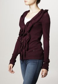 Bordowy kardigan z falbankowym zdobieniem na przodzie, zapinany na guziki, z prążkowanymi mankietami i dołem. Wykonany z miękkiego materiału, o luźnym kroju.