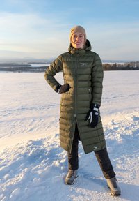 Lång, olivgrön puffjacka med dragkedja, elastiska ärmslut och en logotyp. Kombineras med svarta byxor och grå stövlar. Snöigt bakgrund.