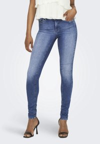 Femme portant un jean skinny bleu, un top blanc sans manches à volants et des sandales noires à talons hauts, debout avec une main dans la poche.