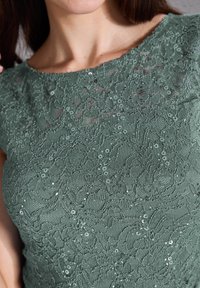 Grünes Spitzenoberteil mit floralem Muster und Pailletten. Der Rundhalsausschnitt und die kurzen Ärmel betonen das strukturierte Material.
