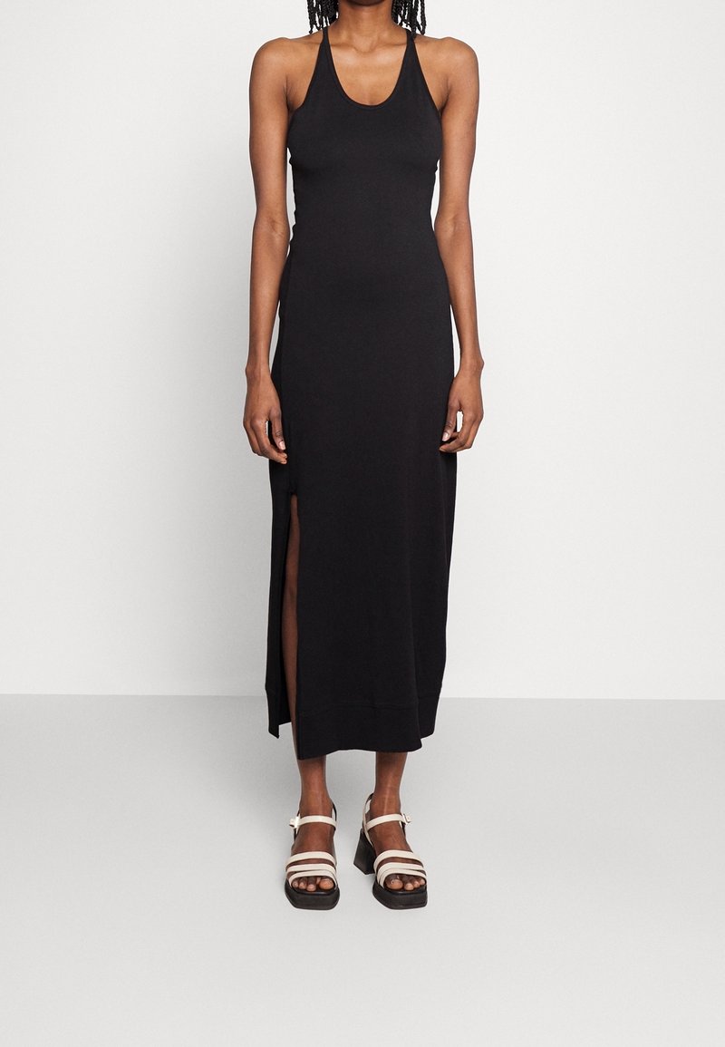 Femme portant une robe maxi noire sans manches avec une fente latérale et des sandales compensées blanches, debout devant un fond blanc uni.