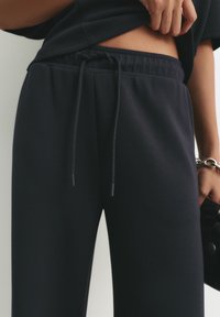 Personne portant un pantalon de survêtement noir avec une taille à cordon, levant légèrement un t-shirt noir pour révéler le bas du torse, tenant un objet noir avec un bracelet en chaîne.