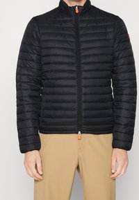 Schwarze gesteppte Daunenjacke mit hohem Kragen, Vorderreißverschluss und orangefarbenem Reißverschlussanhänger. Glatte Textur mit horizontalem Steppdetails.