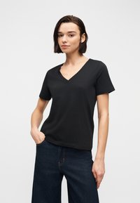 Νεαρό άτομο με κοντά σκούρα μαλλιά, φοράει μαύρο T-shirt με V λαιμόκοψη και σκούρο μπλε τζιν, στέκεται με το ένα χέρι στην τσέπη σε γκρι φόντο.