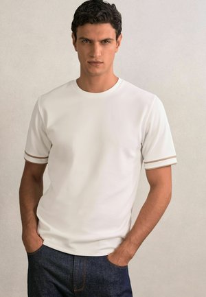 Tricou alb cu mâneci scurte, din material texturat, cu decolteu rotund și dungi accent maro la mansete, asociat cu blugi din denim bleumarin.