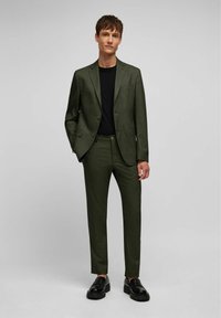 Costume à motifs verts avec une coupe ajustée. Le blazer présente des revers en notch et deux boutons ; associé à un pantalon sur mesure et des chaussures noires.