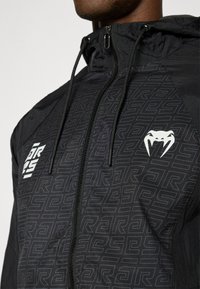 Μαύρο μπουφάν με κουκούλα και γεωμετρικό σχέδιο, με φερμουάρ μπροστά, κορδόνια και λευκές λογοτυπικές λεπτομέρειες στο στήθος και  τον ώμο.