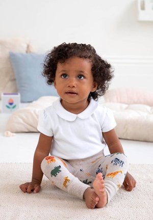 Weißes Kurzarmhemd mit Kragen, kombiniert mit beigen Leggings mit bunten Tiermustern. Kind sitzt auf einem weichen Teppich.