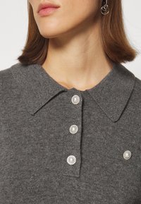 Femme portant un polo en maille grise avec des boutons décoratifs en perles et argent, et un bouton assorti en perle sur une petite poche poitrine.