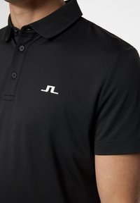 Mustapaita, joka on valmistettu teksturoituneesta kankaasta. Sisältää normaalin kauluksen, kolme nappia ja pienen valkoisen logon vasemmassa rinnassa.