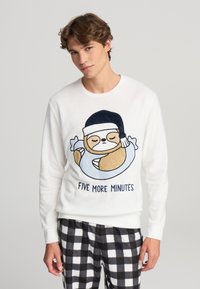 Maglione bianco con un bradipo dei cartoni animati in un tubo blu e la scritta “ANCHE CINQUE MINUTI.” Abbinato a pantaloni da pigiama a quadri bianchi e neri.
