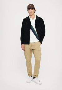 Black corduroy jacket, white polo with green and navy stripes, khaki cargo pants, and white sneakers. Brown cap completes the outfit.