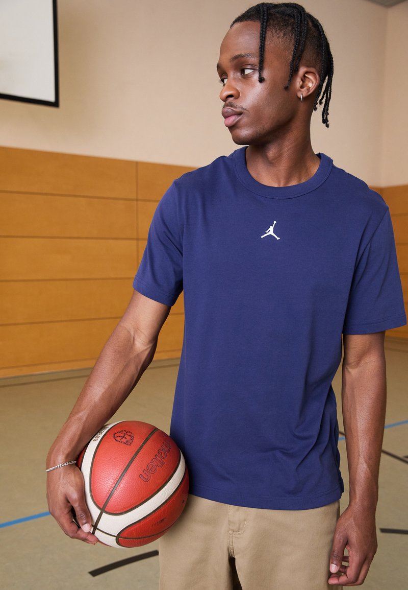 Mørkeblå t-shirt med korte ærmer og logo i hvidt jumpman, båret med beige bukser, mens der holdes en rød og hvid basketball i et træningslokale.
