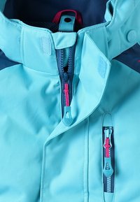 Veste imperméable turquoise avec des accents bleu marine, dotée d'un col haut, de plusieurs zippers en rose et aqua, et d'un tissu texturé.