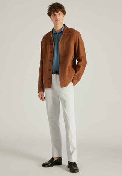 Braune Wildlederjacke mit Reverskragen, geknöpftem Vorderteil und zwei Brusttaschen, über einem petrolfarbenen Poloshirt und cremefarbenen Hosen. Schwarze Slipper.