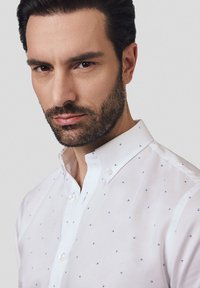 Biała koszula na guziki z małymi niebieskimi kropkami, klasyczny kołnierzyk i dopasowany krój. Materiał ma gładką fakturę i subtelny blask.