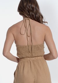 Top halter beige con busto smock e laccio regolabile al collo. Il tessuto appare morbido e leggero, adatto per il clima caldo.