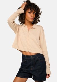 Un pull court beige clair à boutons avec des manches longues, associé à un short en denim foncé. Les détails notables comprennent un col et des boutons à l'avant.