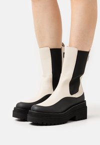 Botas de medio pantorrilla bicolor en blanco y negro con suelas gruesas, llevadas sobre piernas descubiertas contra un fondo neutro.