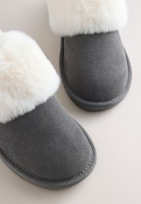 Chaussons en daim gris avec un bord en fausse fourrure blanche moelleuse, un bout arrondi et des détails cousus le long du bord. Texture lisse dans l'ensemble.