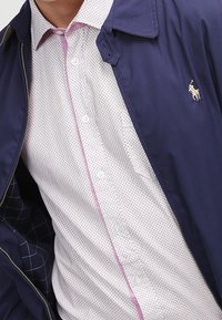 Marine lichtgewicht jack met een rits, voorzien van een kraag en een beigekleurig shirt met patroon eronder, aangevuld met roze knopen en stiksels.