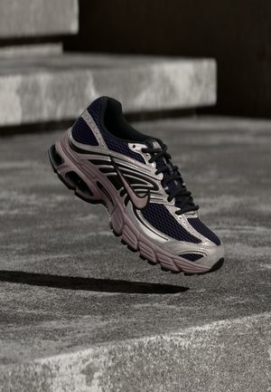 Nike løpesko i svart og grått med rosa detaljer, svevende over en betongflate og trinn i en skyggelagt utendørs setting.