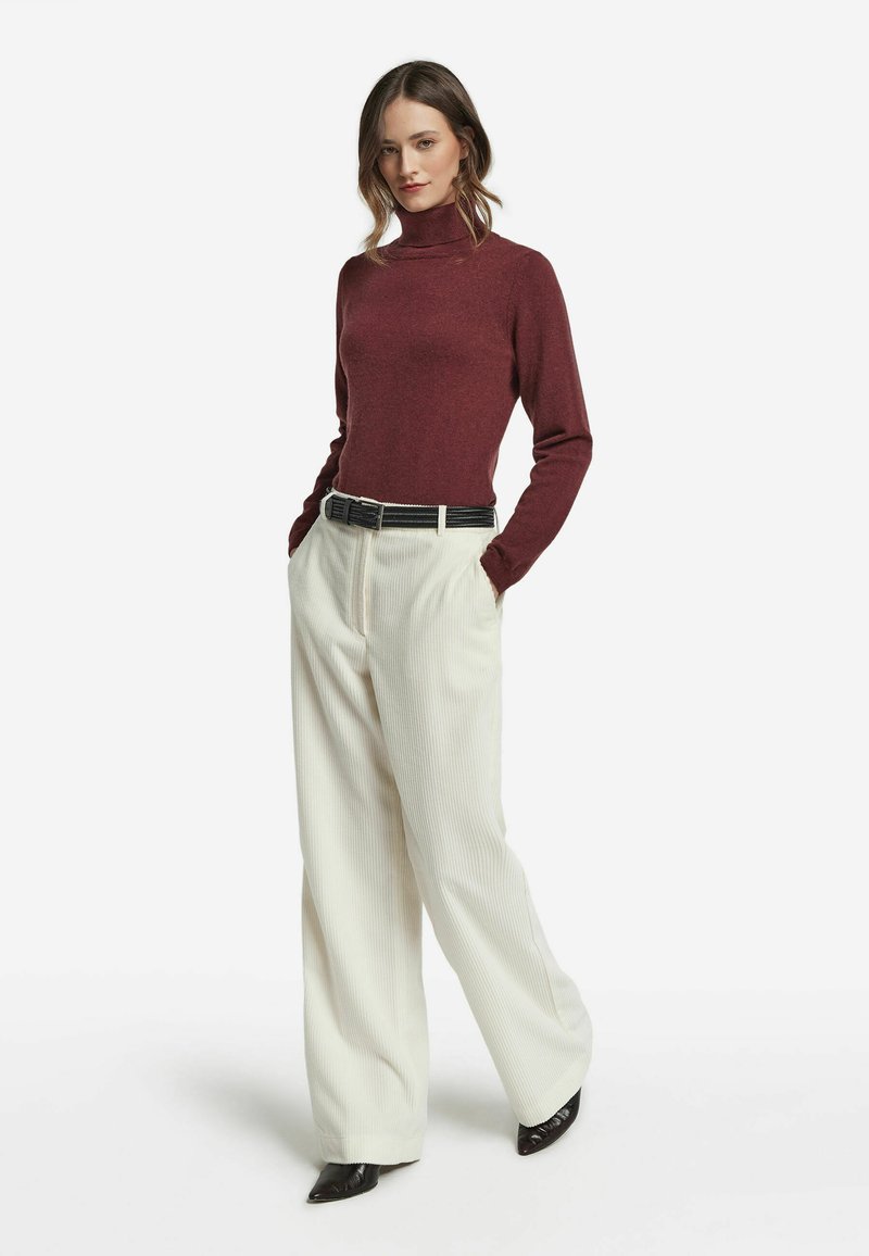Bordeauxfarbener Rollkragenpullover, weite cremfärbige Cordhose, schwarzer Gürtel und spitze schwarze Pumps. Glatte Textur, lässige Passform und vertikale Rippungen.