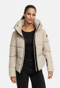Femme portant une doudoune beige avec un col montant, un haut à manches longues noir et un pantalon noir, se tenant devant un fond blanc.