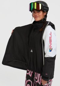 Zwart-witte jas met een ritssluiting aan de voorkant, fleece voering, logodetails en roze accenten, gecombineerd met kleurrijke gestreepte broeken.