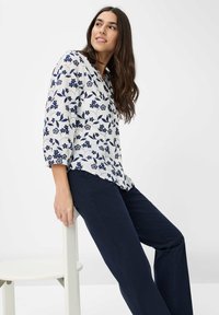 Hvid bluse med marineblåt blomstermønster, v-hals, trekvartærmer, parret med marineblå bukser. Model siddende på en hvid stol.