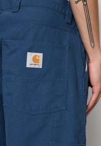 Siniset Carhartt-housut, joissa on suuri takatasku ja logotarra, yllä henkilöllä, jolla on miekka-tatuointi kyynärvarressa.