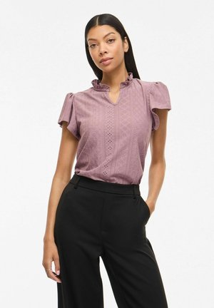 Kobieta ubrana w bluzkę z krótkim rękawem z różowej koronkowej tkaniny z falbaną przy kołnierzu, wpuszczoną w czarne spodnie z wysokim stanem, stojąca z jedną ręką w kieszeni.