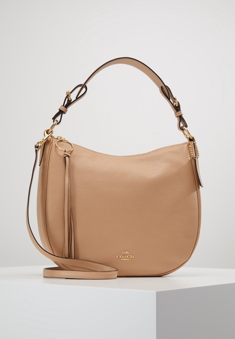 Beige leather shoulder bag with a smooth texture, curved shape, detachable shoulder strap, gold-tone hardware, and tassel detail.