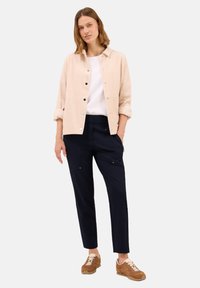Donna che indossa una giacca di velluto a coste beige sopra una camicia bianca, pantaloni blu con tasche e sneaker marroni, in piedi con una mano in tasca.