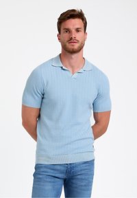 Gaiši zils adīts polo krekls ar īsām piedurknēm, ribotu tekstūru, divu pogu slēgšanu un kontrastējošu apkakli. Valkāts kopā ar ziliem džinsiem.